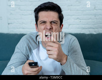 Stile di vita Ritratto di giovane uomo sensazione spaventato e scioccato rendendo la paura, ansia gesti mentre guardano la televisione tenendo premuto il comando remoto. In orrore un Foto Stock