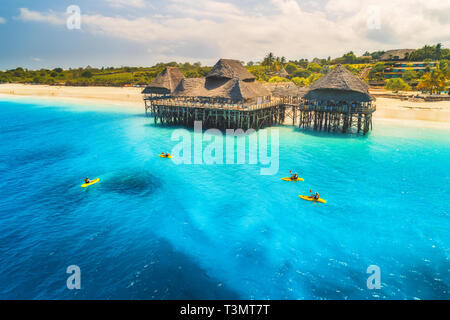 Vista aerea del bellissimo hotel e persone in kayak al tramonto in estate. Zanzibar, Africa. Vista dall'alto. Paesaggio con hotel in legno sul mare, azure wat Foto Stock