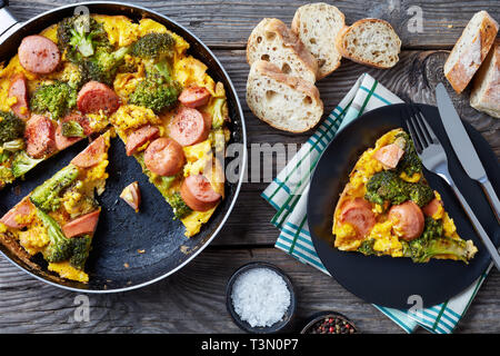 Frittata con broccoli e salsicce in una padella e servita su una piastra su un vecchio grigio a tavola in legno rustico con croccanti fette integrali baguett francese Foto Stock