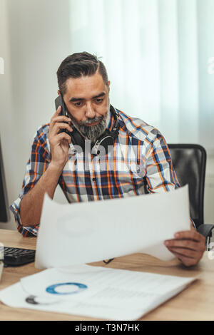 Un imprenditore di successo, con cuffie intorno al suo collo, sta parlando su uno smartphone e il controllo dei documenti cartacei in ufficio. Foto Stock