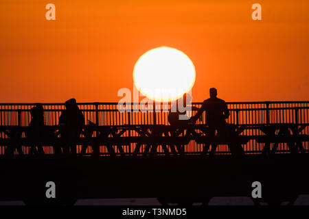 Aberystwyth, UK. Decimo Apr, 2019. Regno Unito: meteo alla fine di una giornata ininterrotte blu del cielo e il caldo sole primaverile, il sole tramonta spettacolarmente dietro le sagome delle persone gustando un drink sul mare dal molo di Aberystwyth su Cardigan Bay costa del Galles occidentale. Credito: keith morris/Alamy Live News Foto Stock