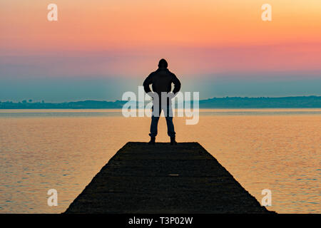 L'uomo stagliano permanente sulla scogliera che si estende fino al mare di sunrise Foto Stock