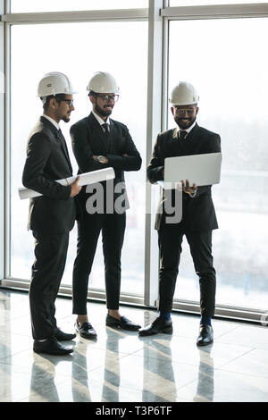 Dettaglio di tre diverse sorridente business persone che indossano caschi e di stare all'aperto con edificio in background. Concetto di architetti. Foto Stock