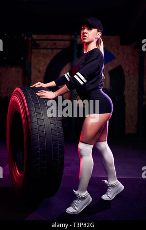 Montare attraente medioevo atleta donna in posa con un enorme pneumatico in palestra. Montare cross donna con grandi pneumatici. Foto Stock