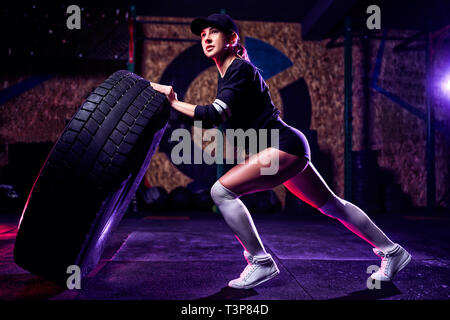 Montare attraente atleta donna che lavora fuori con un enorme pneumatico, la rotazione e il capovolgimento in palestra. Montare cross donna esercitando con pneumatici di grandi dimensioni. Foto Stock