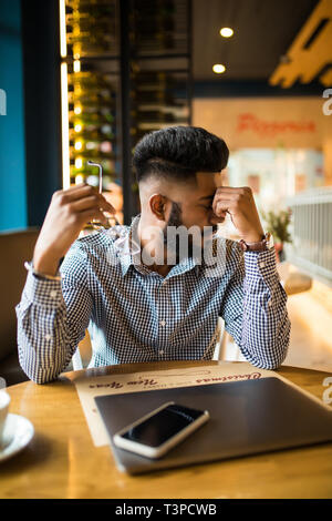 Uomo indiano utilizza lo smartphone e il computer portatile in cafe sotto stress e di preparazione per gli esami Foto Stock