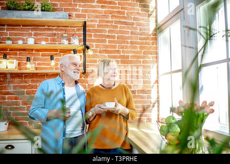 Coppia senior avente un tempo di caffè, in piedi insieme vicino alla finestra sulla cucina a casa Foto Stock