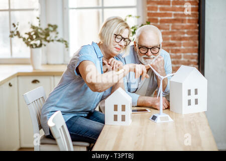Coppia senior dreaming about proprietà moderna e di energie alternative, seduta con modelli della casa e toy turbina eolica a casa Foto Stock