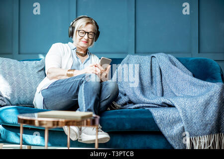 Senior donna vestita casualmente ascoltando la musica con le cuffie e telefono sul lettino a casa Foto Stock
