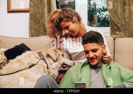Sorridente giovane coppia eterosessuale insieme sul divano con il loro cane utilizza lo smartphone Foto Stock