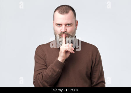 Uomo maturo con barba in maglietta marrone compiendo il gesto del silenzio Foto Stock