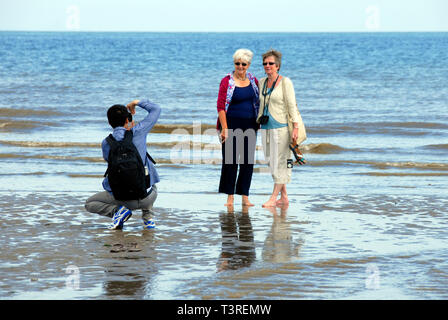 Giovane uomo prendendo fotografia di due anziane signore pagaiando sulla spiaggia Foto Stock