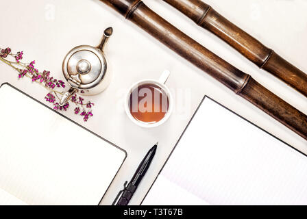 Composizione piana cup con tè, bollitore d'argento, pastiglie, penna, viola fiori secchi, steli di bambù Foto Stock