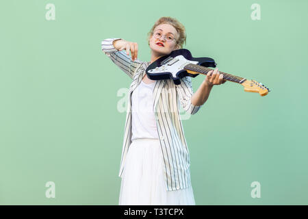 Ritratto di giovane donna bionda in maglia bianca, gonna e camicia a righe con occhiali in piedi e tenere la chitarra elettrica e cercando. prigioniero al coperto Foto Stock