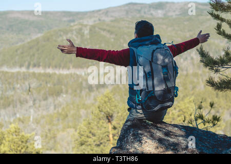 Un maschio di turista escursionista con un zaino grande sorge su una scogliera pietra davanti di una verde vallata con braccia aperte. Il concetto di libertà in tr Foto Stock