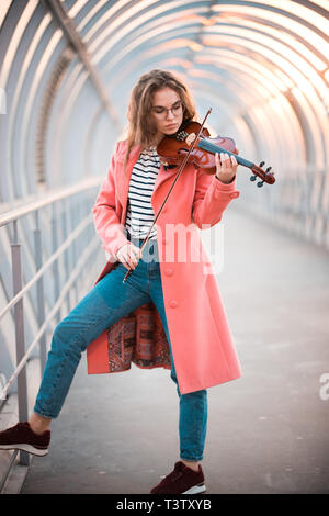 Giovane donna in cappotto rosa in piedi sul passaggio di overhead suona il violino Foto Stock