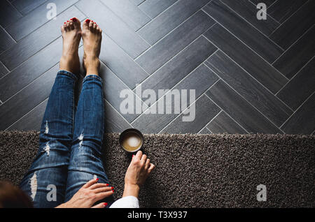 Donna con le mani unghie rosse azienda tazza di caffè sulle ginocchia wering jeans blu Foto Stock