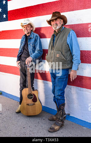 Due vecchi cowboy con cappelli da cowboy di stare in piedi insieme nella parte anteriore di un rosso e pareti bianche e uno di molti è in possesso di una chitarra. Foto Stock