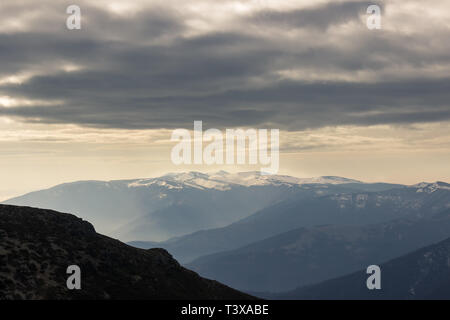Moody, drammatico cielo sopra appuntite, distante, impressionante vertice Kom in Bulgaria coperti di neve e nebbioso e in primo piano gli strati di montagna Foto Stock