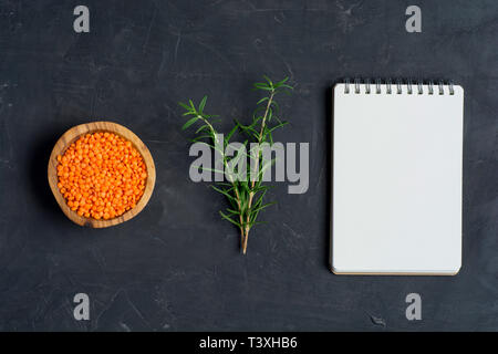 Di lenticchie rosse nella ciotola di legno con rosmarino e blocco note su un calcestruzzo scuro dello sfondo. Appartamento laici o vista dall'alto. Foto Stock