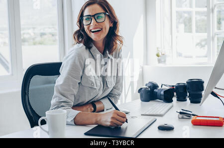 Allegro fotografo femmina lavorando alla sua scrivania in ufficio. Donna con grafica digitale tavoletta e penna di disegno che guarda lontano e sorridente. Foto Stock
