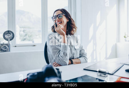 Femmina professionista creativo seduto alla sua scrivania che guarda lontano e sorridente. Donna fotografo nel suo ufficio. Foto Stock