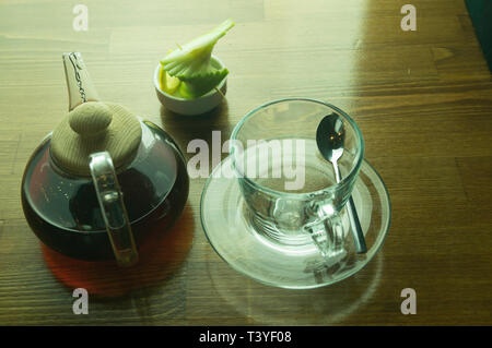 Un bicchiere di infuso di tè e un bicchiere con il limone in una teiera di vetro collocato su un tavolo marrone Foto Stock