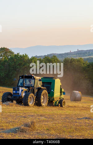 Giugno 18, 2018 MONTEGABBIONE, Umbria, Italia: un fieno imballatrice a lavorare in un campo come il sole tramonta. Foto Stock