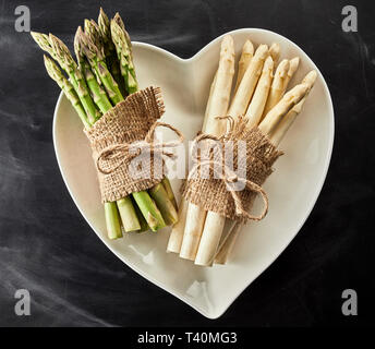 A forma di cuore ad ciotola con due fasci di colore bianco e verde di asparagi freschi spears legato con una corda e hessian su un sfondo di ardesia in formato quadrato Foto Stock