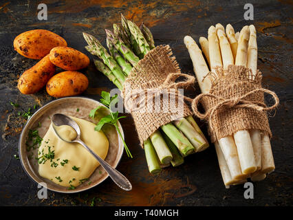 Crudo fresco asparagi spears legate in fasci con la maionese in un recipiente laterale e patate novelle su un rustico sfondo metallico visto dal di sopra Foto Stock