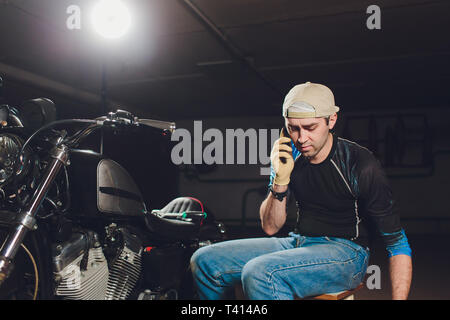 Uomo che fissa la bicicletta. Fiducioso giovane uomo che ripara moto presso il suo garage. prendendo gli ordini tramite telefono Foto Stock