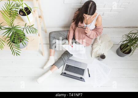 Una giovane donna felice si siede sul pavimento in un luminoso appartamento o di un ufficio interno e lavora su un portatile, freelancer ragazza al lavoro, il concetto di distanza Foto Stock