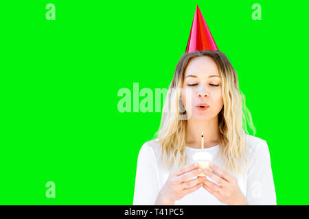 Una millenaria donna con capelli biondi celebra un compleanno con una festa di compleanno cupcake e compleanno hat. Brucia la candela Foto Stock