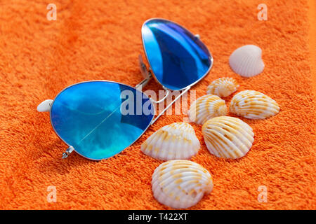 Moda sunglases vicino a conchiglie sulla spiaggia arancione asciugamano. Fotografia concettuale suggerendo le vacanze estive e il tempo per il relax e la pastella di carica Foto Stock