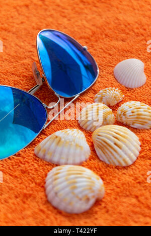 Moda sunglases vicino a conchiglie sulla spiaggia arancione asciugamano. Fotografia concettuale suggerendo le vacanze estive e il tempo per il relax e la pastella di carica Foto Stock