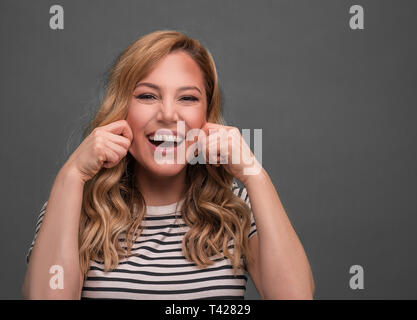 Una giovane donna si allunga la sua bocca in un ipocrita sorriso contro uno sfondo grigio. Sorriso. Foto Stock