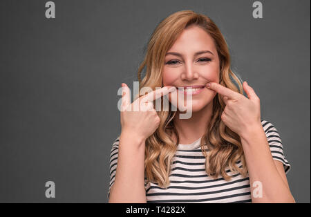 Una giovane donna si allunga la sua bocca in un ipocrita sorriso contro uno sfondo grigio. Sorriso. Foto Stock