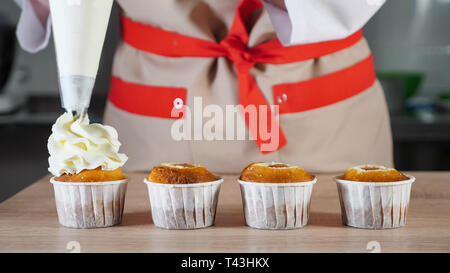 Pastry chef decora i tortini in bicchieri di carta bianco crema con sacca di tela da pasticceria. Le mani di close-up. vista frontale Foto Stock