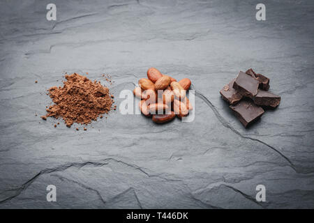 Organici di chicchi di cacao, la polvere di cacao e cioccolato su una pietra nera dello sfondo. Composizione orizzontale Foto Stock