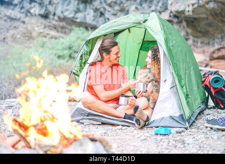 Coppia felice degli escursionisti con il loro cane camping con tenda intorno montagne di roccia accanto al fuoco - Persone rilassante in un accampamento di bere il tè caldo Foto Stock