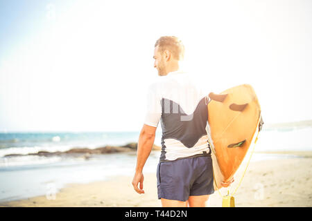 Giovani surfer tenendo la sua tavola da surf guardando le onde per il surf - uomo bello permanente sulla spiaggia al tramonto la formazione per il surf Foto Stock