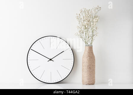 Vista frontale tavolo rotondo con orologio a parete e fiori in vaso bottiglia su sfondo bianco. Home office il minimo spazio di lavoro scrivania Foto Stock