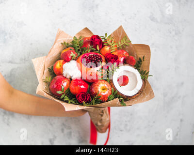 Frutta e bacche bouquet. Mangiare bouquet in mano femmina su sfondo grigio. Melograno, fragola, mele, noci di cocco e le rose fiori, eucalipto. DOF poco profondo. Spazio di copia Foto Stock