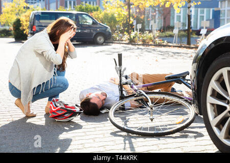 Giovane donna Guardando inconscio ciclista maschio giacente sulla strada dopo un incidente nei pressi di auto Foto Stock