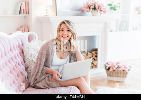 Ritratto di una splendida bionda donna con notebook a lavorare a casa. Donna seduta sul divano rosa in luce interni di lusso con camino e fiori. Donna Foto Stock