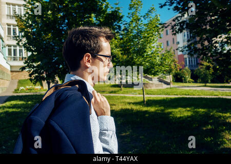 Giovane imprenditore con gli occhiali della sua maglietta è camminare per la strada e la holding acket sulla sua spalla. Visualizza profilo Foto Stock