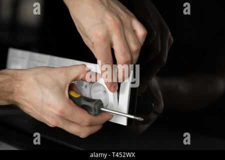 Close-up di un elettricista la mano con un cacciavite smontare un bianco presa elettrica su una nera parete di vetro. Foto Stock