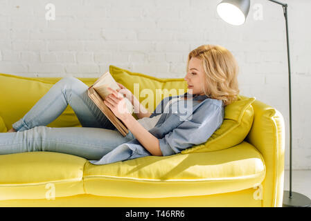 Donna sdraiata sulla accogliente lettino giallo e il libro di lettura Foto Stock