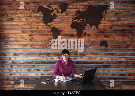Grave donna che lavorano alla sua scrivania in ufficio a prendere appunti come Lei naviga su Internet con un computer portatile al di sotto di una mappa del mondo sulla parete Foto Stock