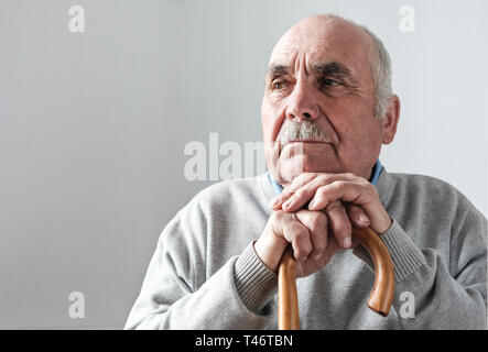 Anziani dai capelli grigi pensionati uomo con bastone seduto appoggiato il mento sulla sommità delle sue mani sulla canna staring a lato nel profondo del pensiero Foto Stock